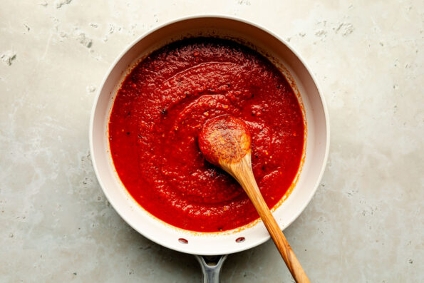 An overhead shot of pasta sauce and a wooden spoon in a white frying pan atop a beige surface.