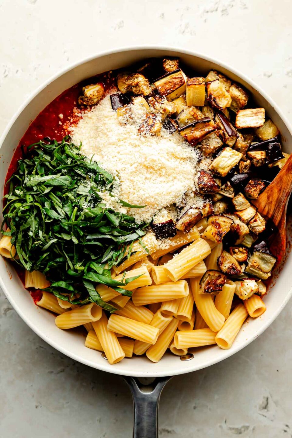 An overhead shot of noodles, chopped basil, roasted eggplant, parmesan and pasta sauce in a white frying pan atop a beige surface.