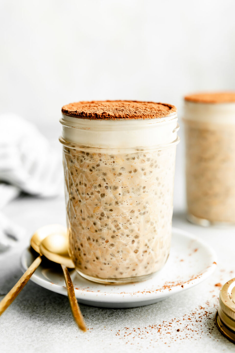 A jar of tiramisu overnight oats topped with espresso powder sits on a small white plate beside two gold spoons atop a light grey surface.