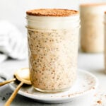 A jar of tiramisu overnight oats topped with espresso powder sits on a small white plate beside two gold spoons atop a light grey surface. Two additional jars of overnight oats and a striped cloth sit in the background.