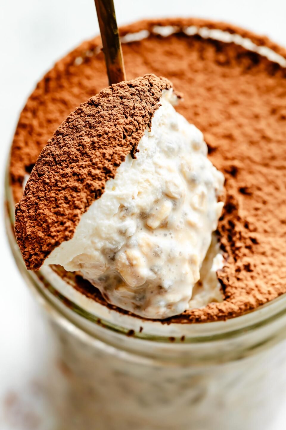 A close-up macro shot of a spoon holding up overnight oats topped with whipped cream and espresso powder over a jar of overnight oats.