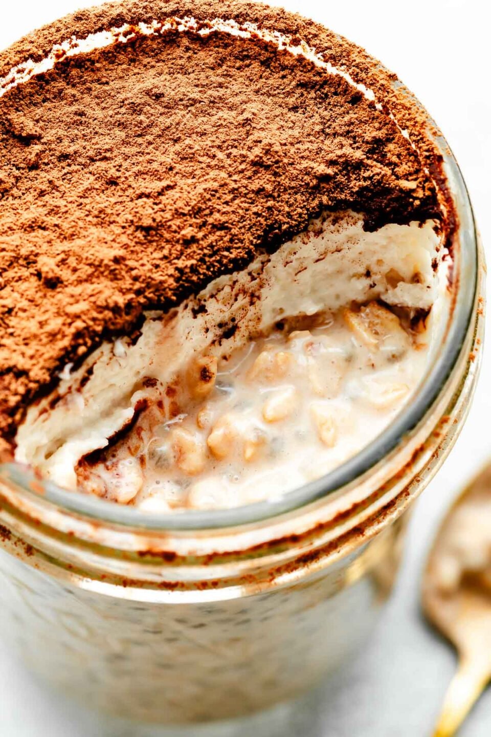 A close-up macro shot of a jar of tiramisu overnight oats with a spoonful taken out. The layers of whipped cream and espresso powder are visible over the oats.