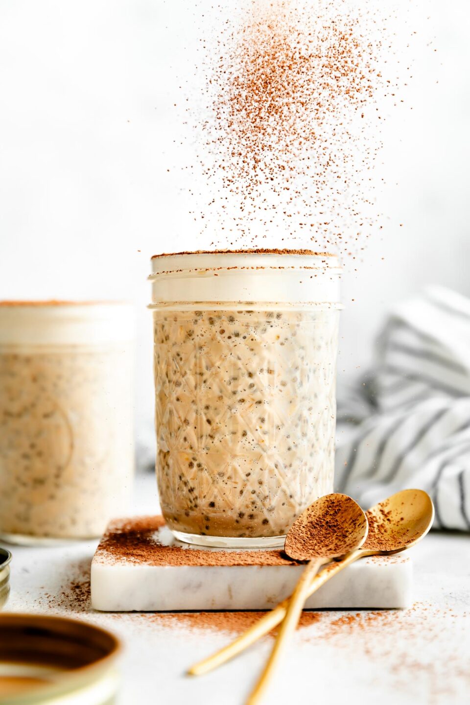 A side shot of a jar of tiramisu overnight oats sitting atop a marble coaster on a white surface. Two gold spoons rest on the coaster, and espresso powder is being sprinkled over the oats. Additional espresso powder sits at the base of the jar. A second jar of oats and a striped towel are seen in the background.