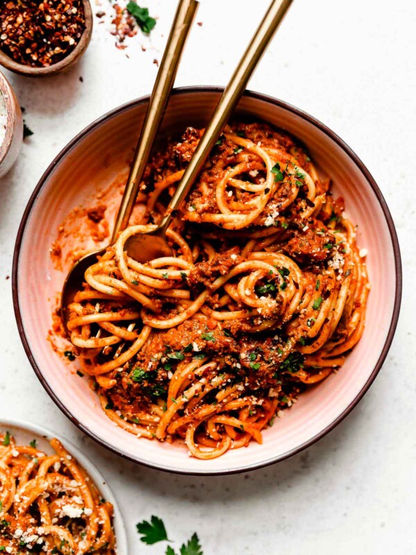 An overhead shot of pasta bolognese in a large white bowl atop a white surface. The bowl sits alongside a second bowl of pasta and dishes of salt and red pepper flakes.