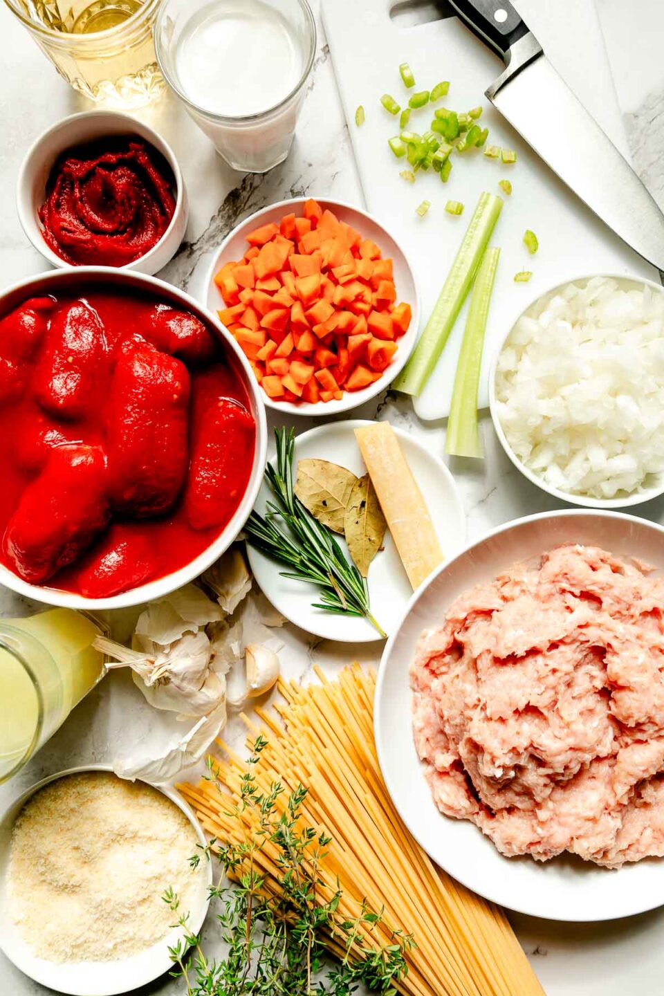 Ingredients are displayed in white dishes on a white marbled surface: ground chicken, dry bucatini, carrots, celery, onion, parmesan, white wine, tomato paste, rosemary, bay leaves, parmesan rind and garlic.