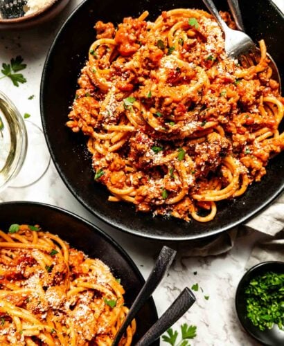 An overhead shot of two black bowls of chicken bolognese pasta atop a white marbled surface. A glass of wine and small bowls of parsley, red pepper flakes and grated parmesan sit beside the bowls.