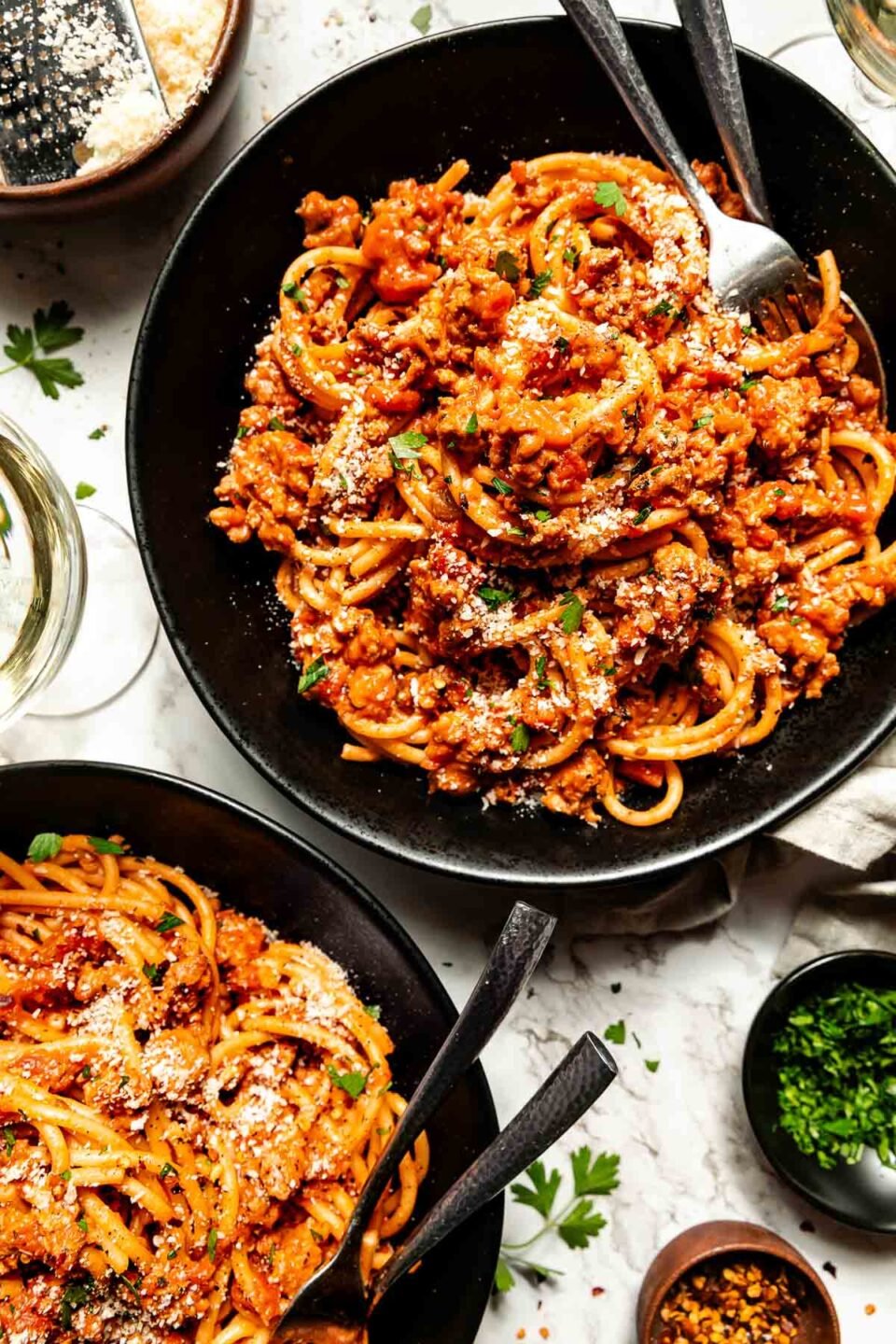 An overhead shot of two black bowls of chicken bolognese pasta atop a white marbled surface. A glass of wine and small bowls of parsley, red pepper flakes and grated parmesan sit beside the bowls.