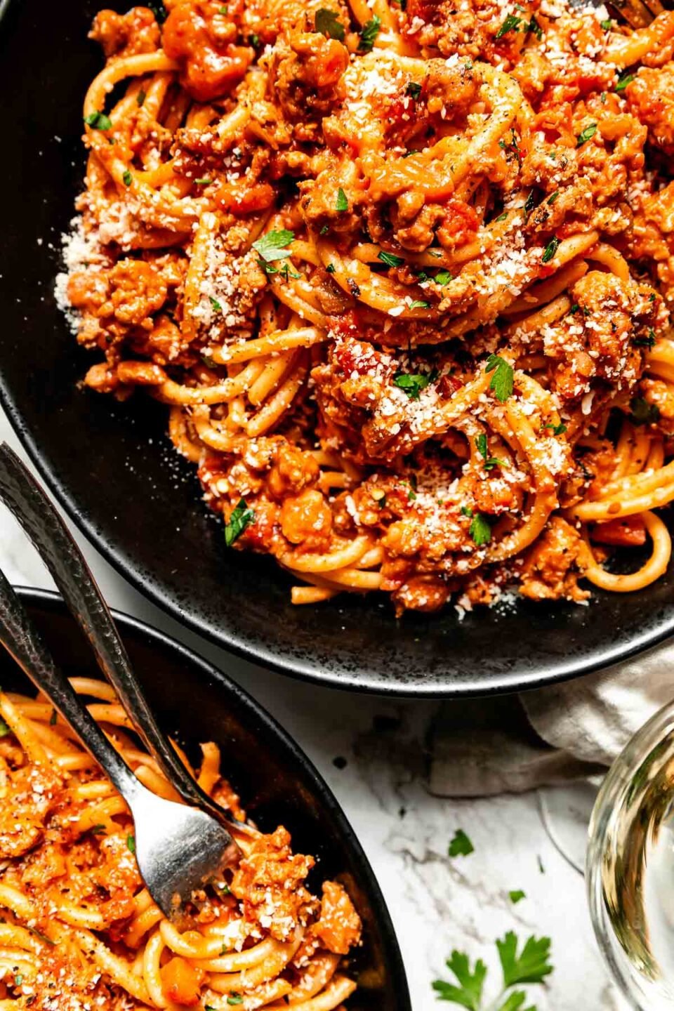 A close-up overhead shot of two black bowls of chicken bolognese pasta beside a glass of white wine atop a white marbled surface.