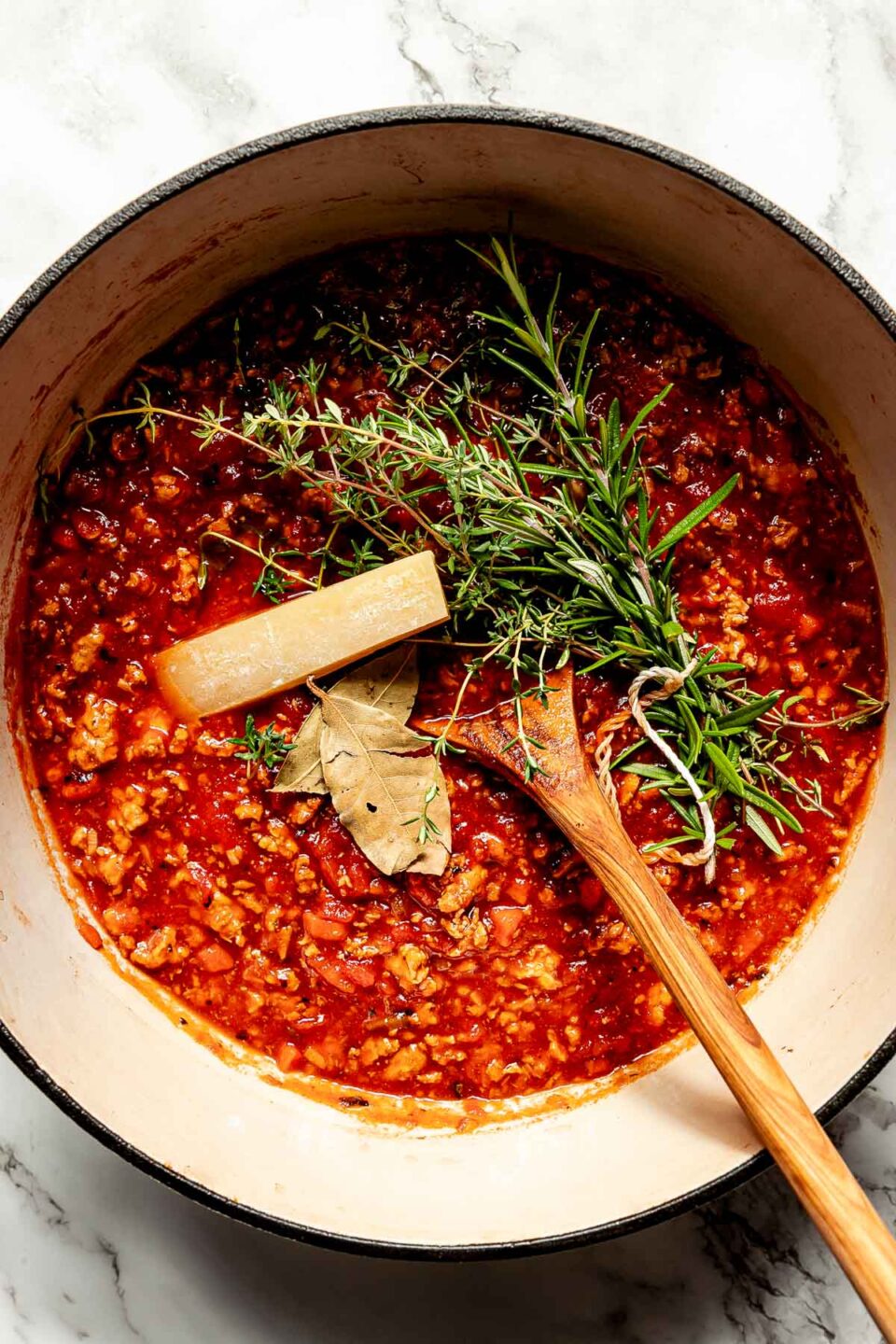 An overhead shot of sauce, a parmesan rind, a tied bundle of herbs, bay leaves and a wooden spoon in a white Dutch oven atop a white marbled surface.