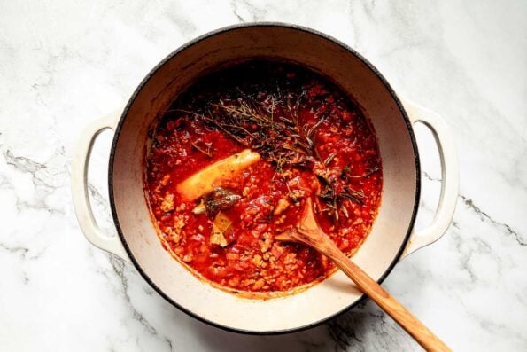 An overhead shot of cooked bolognese sauce with a wooden spoon in a white Dutch oven atop a white marbled surface.