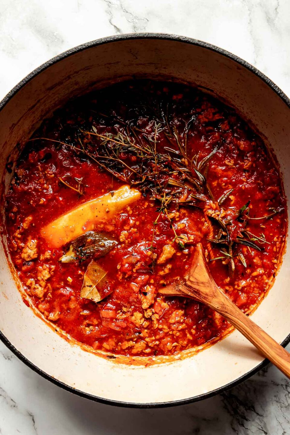An overhead shot of cooked bolognese sauce with a wooden spoon in a white Dutch oven atop a white marbled surface.