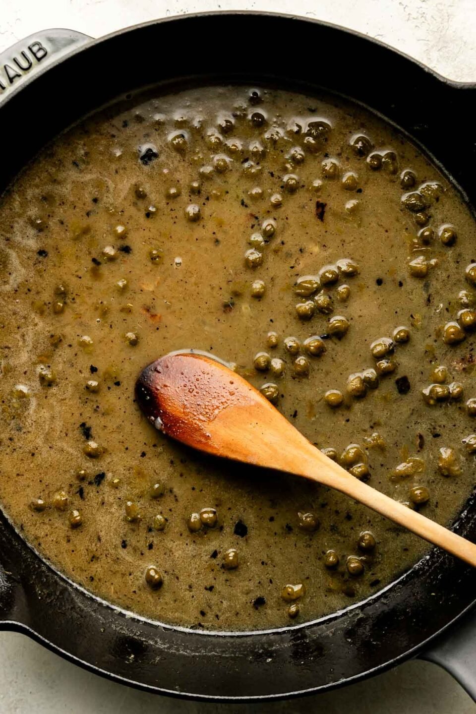 An overhead shot of piccata sauce with a wooden spoon in a black skillet atop a textured off-white surface.