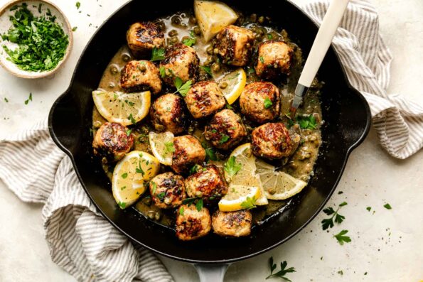 An overhead shot of chicken piccata meatballs garnished with lemon slices and fresh parsley in a black skillet. The skillet sits atop a striped cloth on an off-white surface, and a small bowl of chopped parsley sits beside it.
