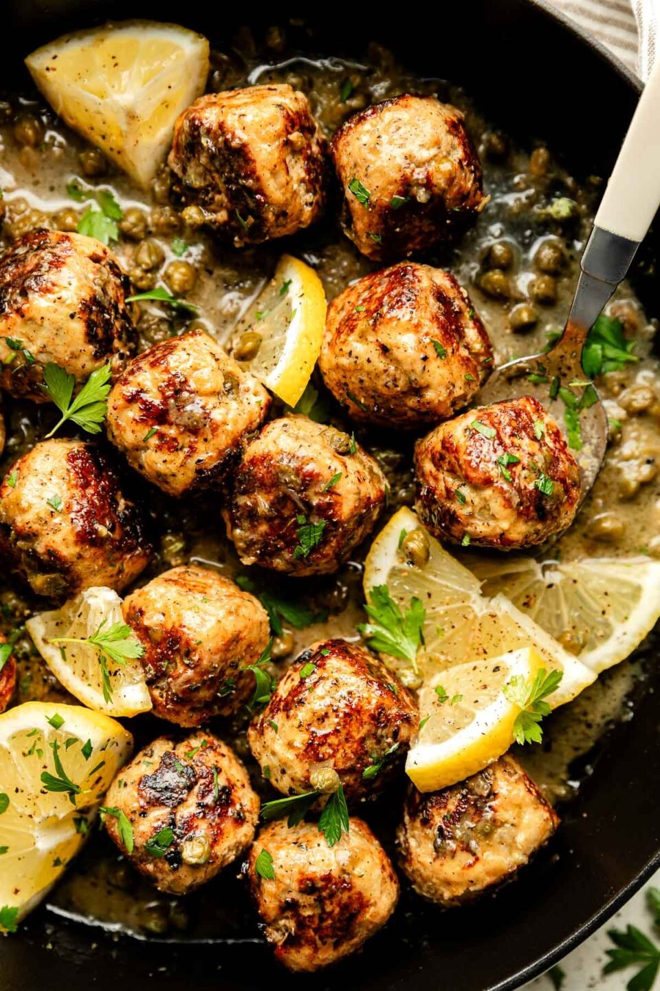 An overhead shot of chicken piccata meatballs garnished with lemon slices and fresh parsley in a black skillet.