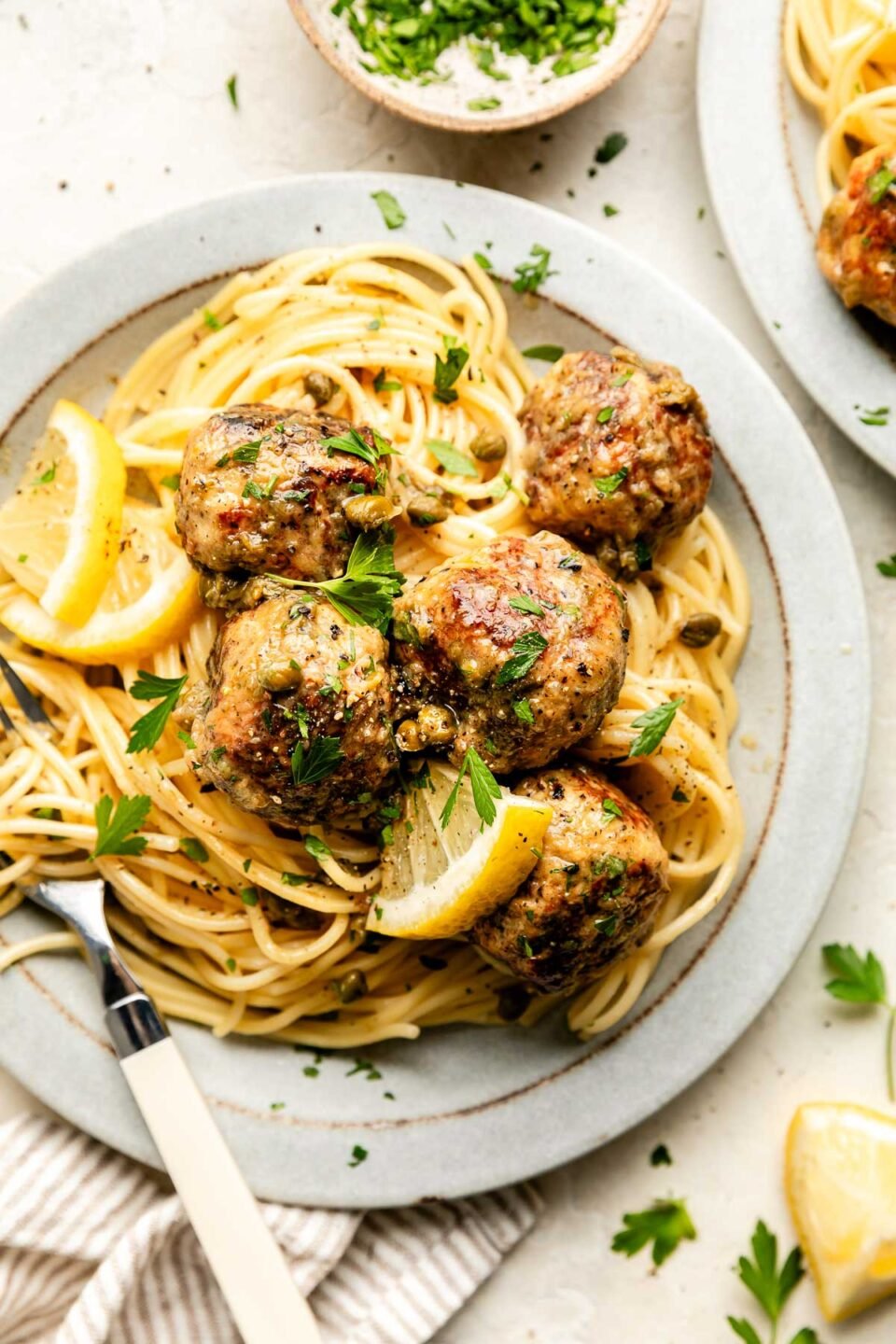 An overhead shot of a serving of chicken piccata meatballs on a bed of spaghetti noodles on a white stoneware plate atop an off-white surface. A second plate sits beside it.