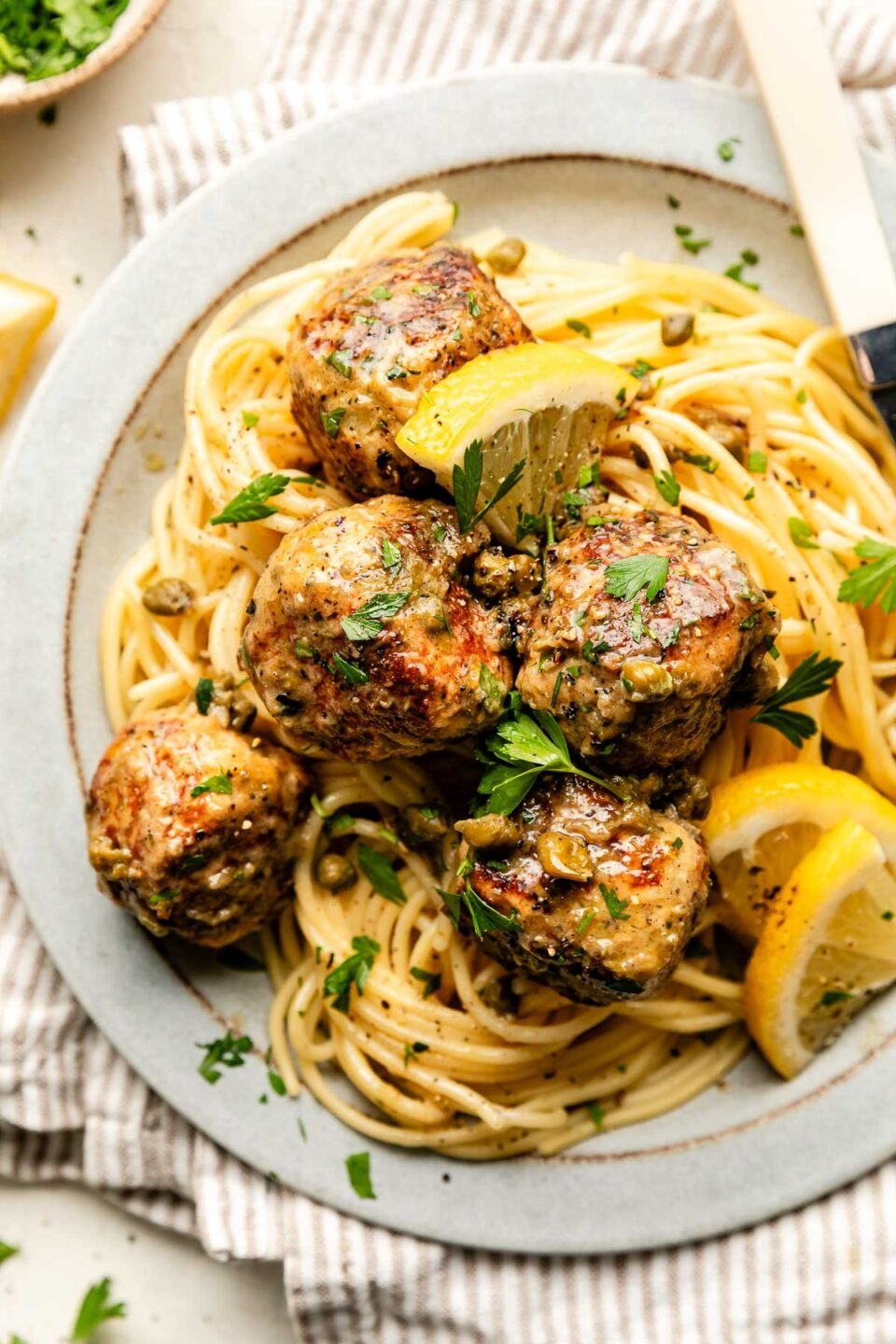 An overhead shot of a serving of chicken piccata meatballs on a bed of spaghetti noodles on a white stoneware plate atop a striped cloth on an off-white surface.