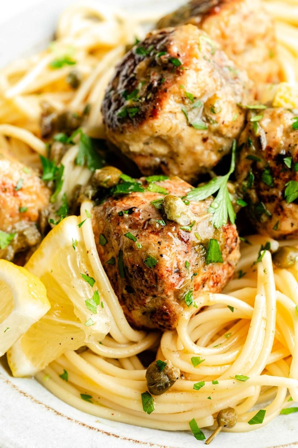 A close-up macro shot of chicken piccata meatballs on a bed of spaghetti, garnished with lemon slices and chopped parsley.