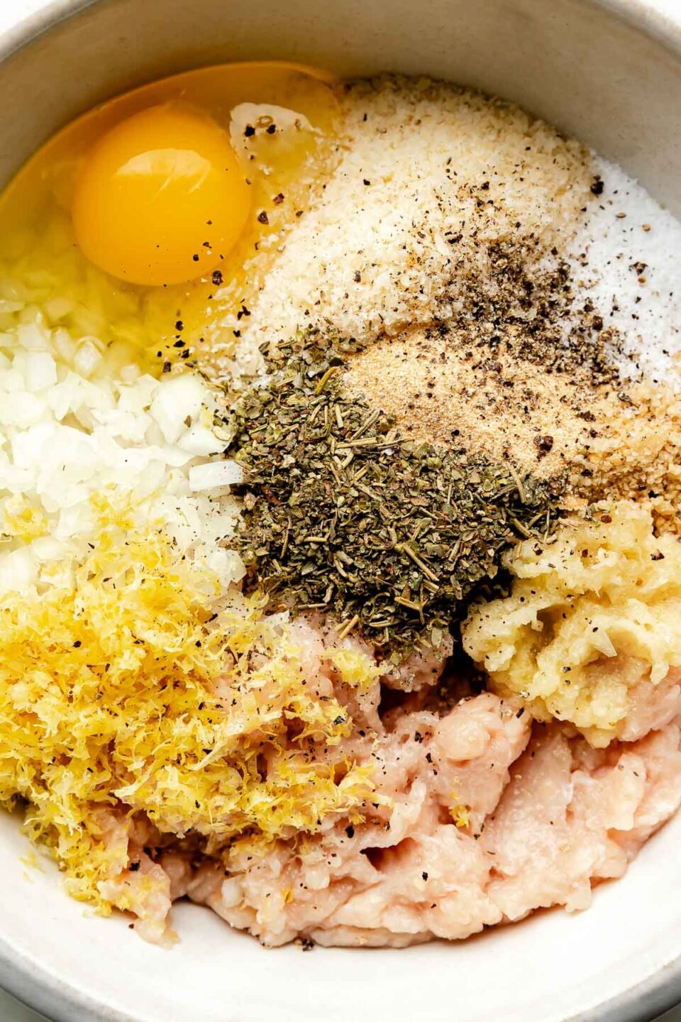An overhead shot of ingredients in a white stoneware bowl: ground chicken, lemon zest and juice, egg, white wine, chicken stock, breadcrumbs, onion, grated parmesan, Italian seasoning and garlic powder.