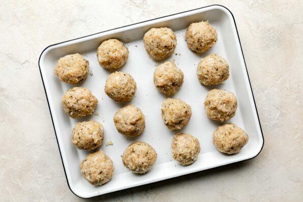 An overhead shot of 16 formed meatballs on a white sheet pan atop an off-white textured surface.