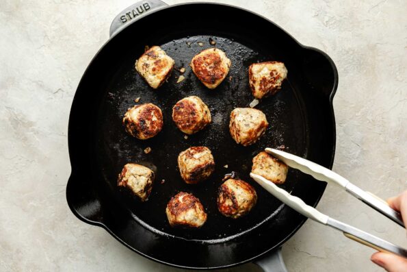 An overhead shot of meatballs being browned in a black cast iron skillet atop a textured off-white surface. A pair of tongs is turning one of the meatballs.
