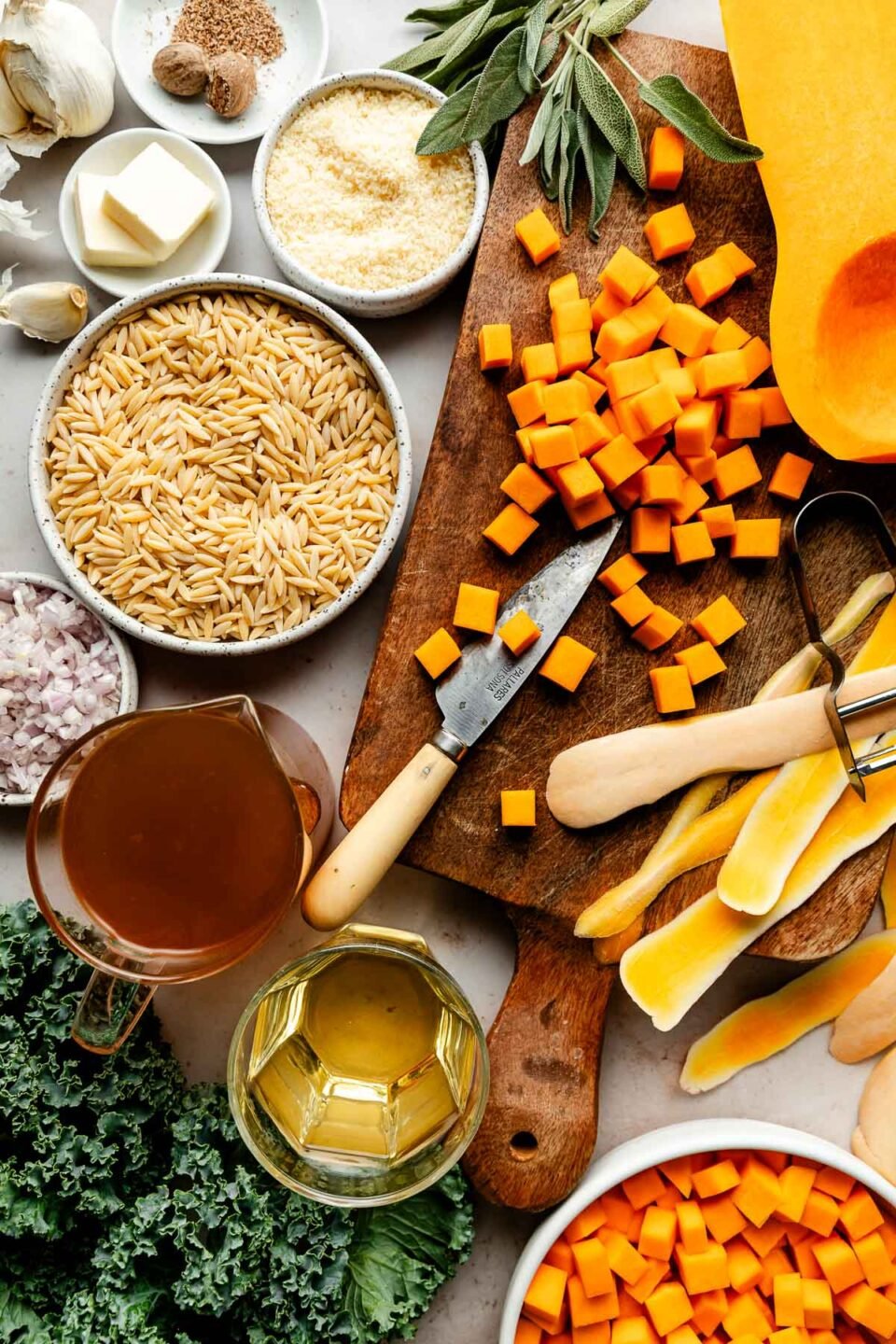 An overhead shot of ingredients displayed in various bowls and on a wooden board on a white surface: diced butternut squash, dry orzo, white wine, broth, shallots, parmesan, butter, garlic, sage, and kale.