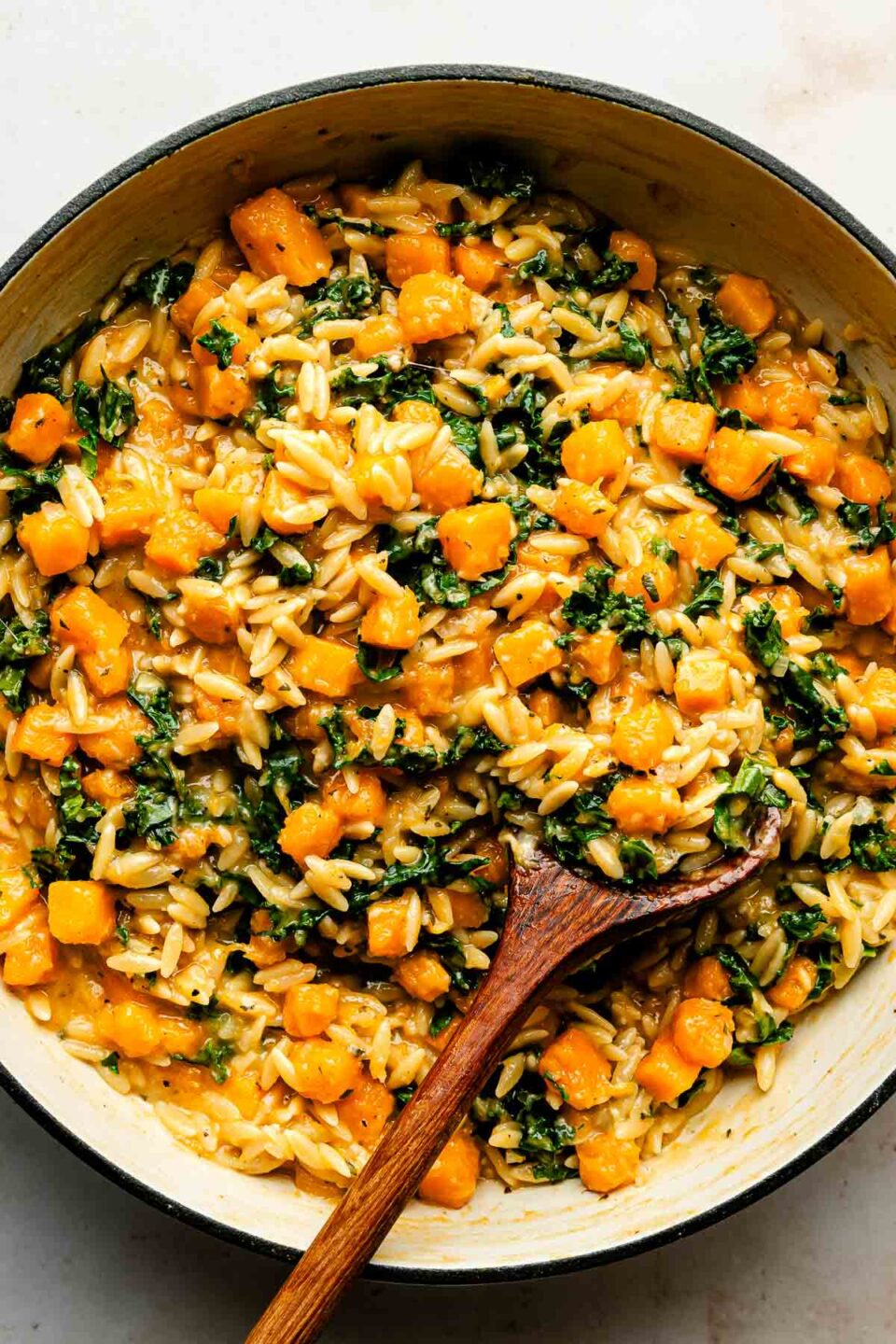 An overhead shot of a wooden spoon resting in a skillet full of cooked squash, kale and orzo atop a textured off-white surface.