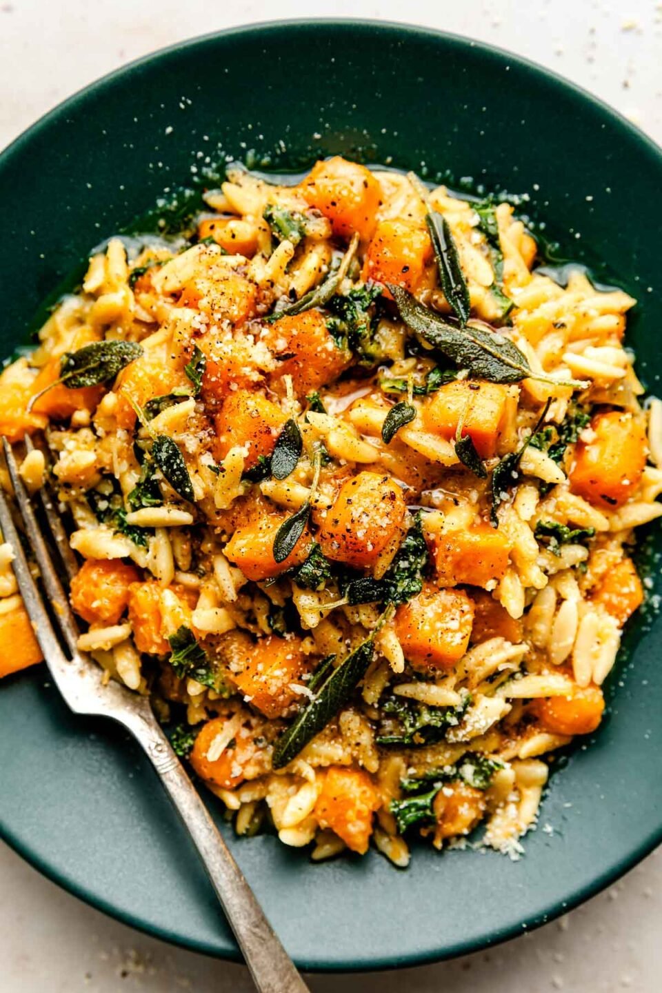 An overhead shot of a serving of butternut squash orzo topped with grated parmesan in a dark green shallow bowl atop an off-white surface.