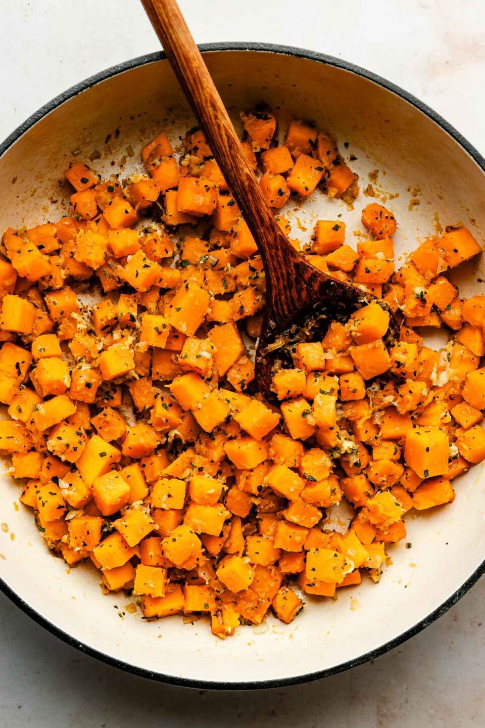 An overhead shot of softened diced squash with a wooden spoon in a white skillet.