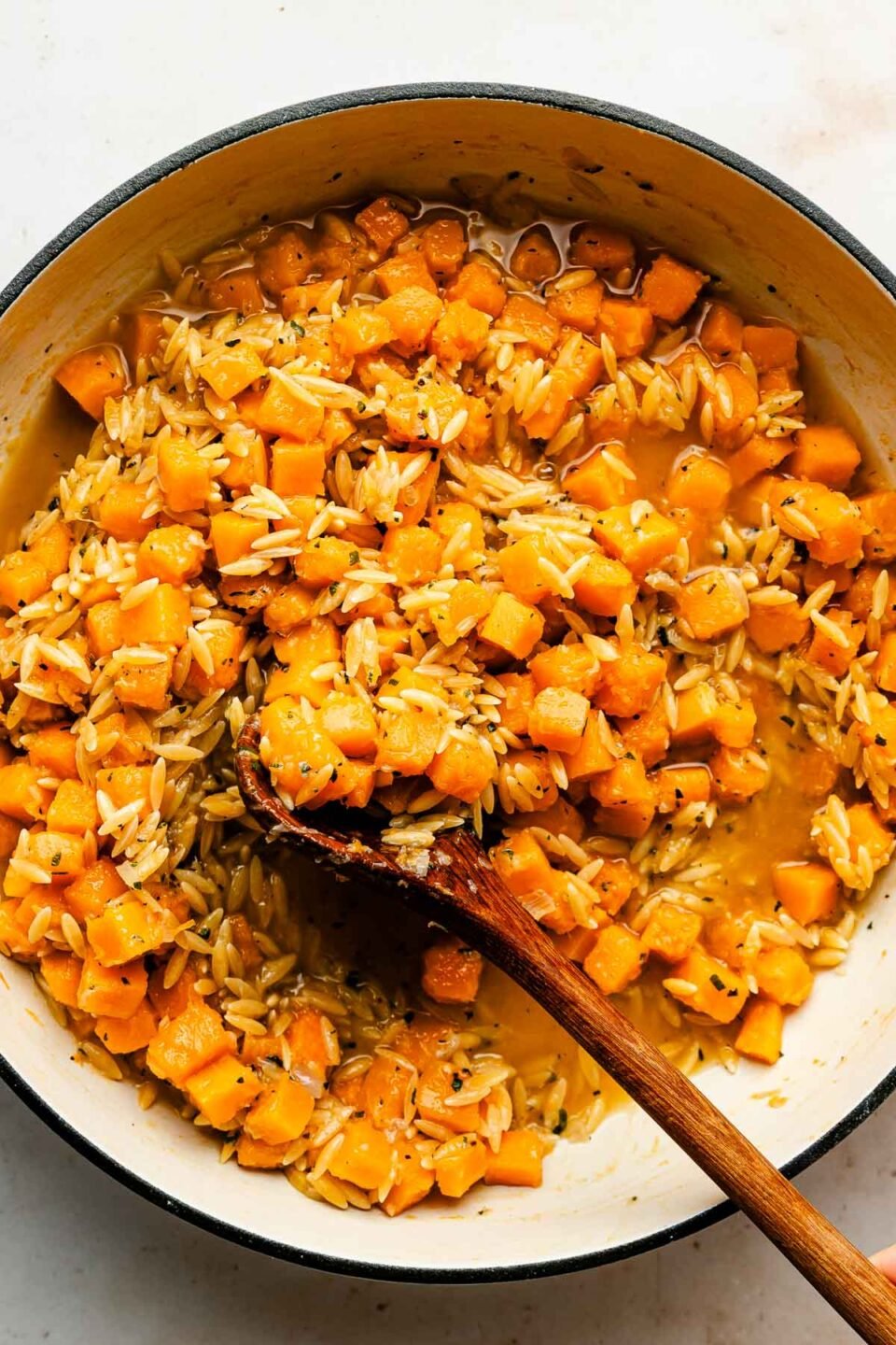 An overhead shot of a wooden spoon stirring squash and orzo in a white skillet.