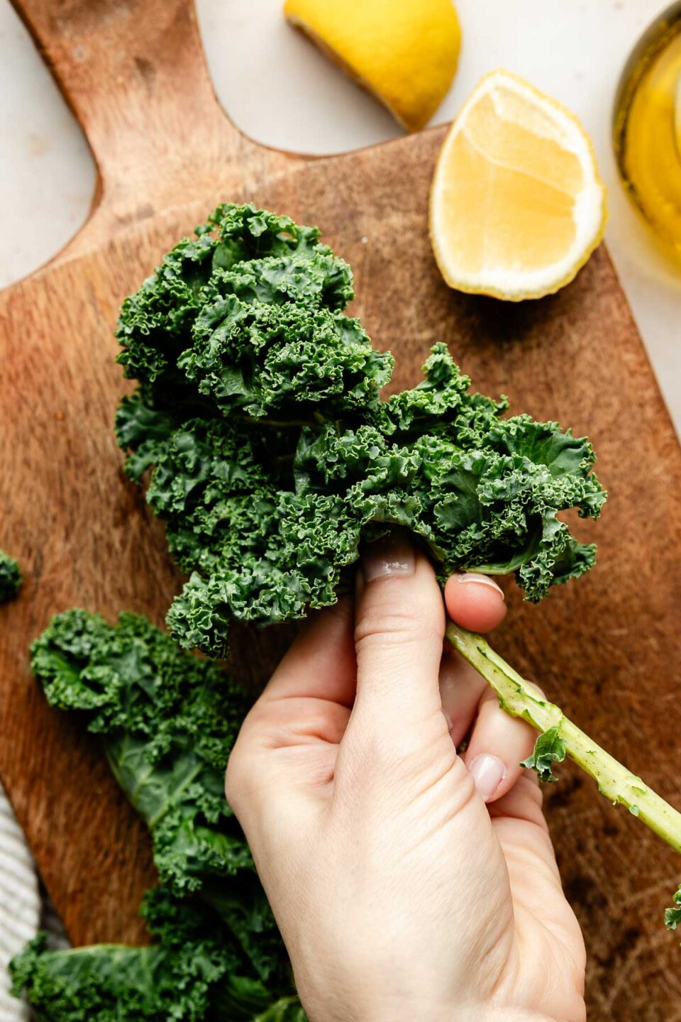 An overhead shot of a woman's hand de-stemming a stalk of kale over a wooden board. Lemon wedges sit on the board and beside it on a white surface.