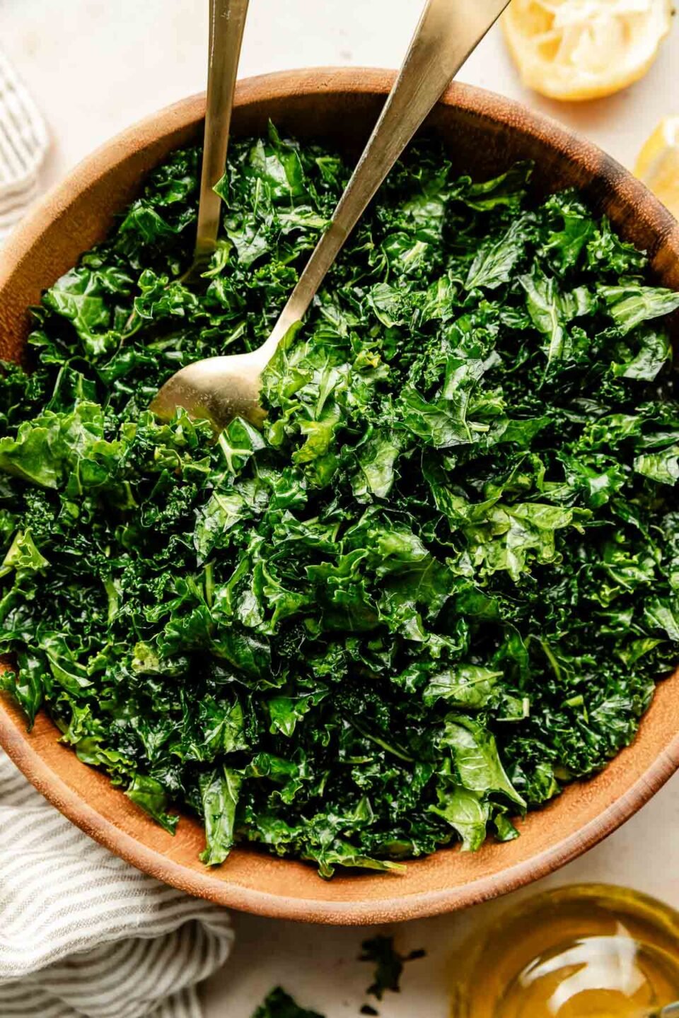 An overhead shot of kale in a large wooden bowl atop an off-white surface. A striped cloth and squeezed lemon wedges sit beside the bowl.