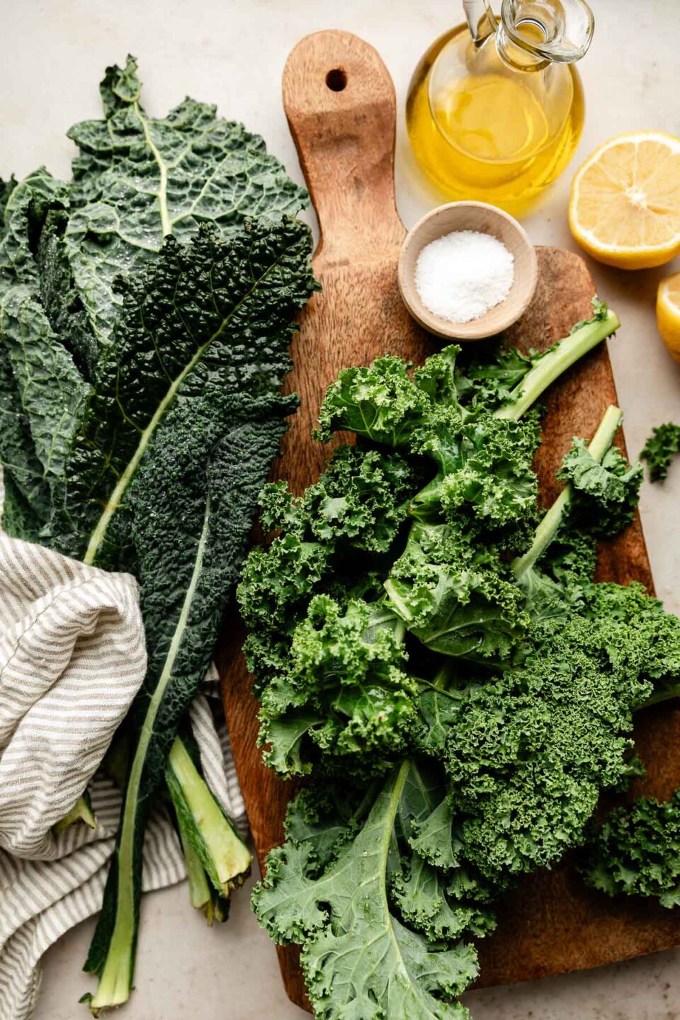 An overhead shot of kale, a jug of olive oil, halved lemons and a small bowl of sea salt on a wooden board and an off-white surface.