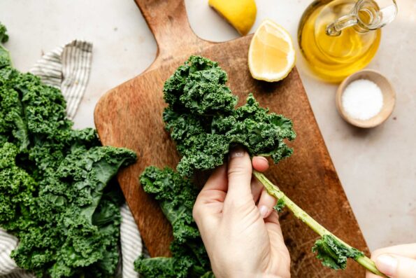 An overhead shot of a woman's hand de-stemming a stalk of kale over a wooden board. Lemon wedges, additional kale, a bowl of sea salt and a jug of olive oil sit on the board and beside it on a white surface.