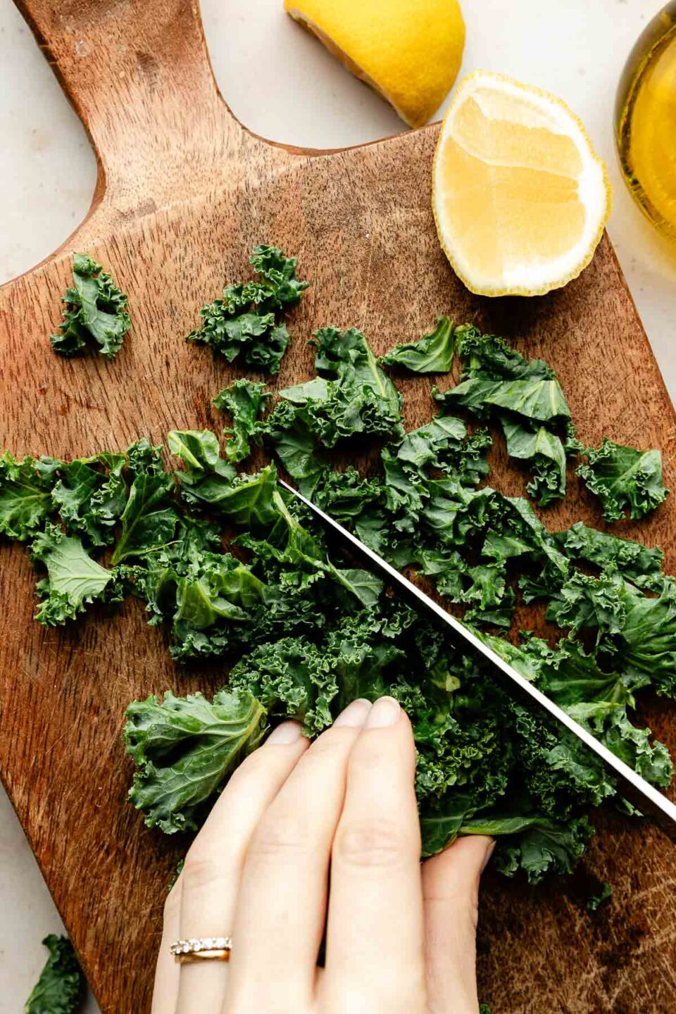 An overhead shot of a woman's hand holding kale and chopping it on a wooden board. Lemon wedges sit beside the kale.