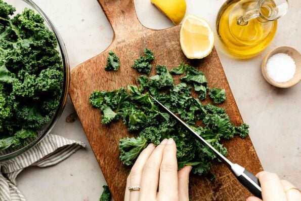An overhead shot of a woman's hand holding kale and chopping it on a wooden board. Lemon wedges, a bowl of chopped kale, salt and a jug of olive oil sit beside the kale.