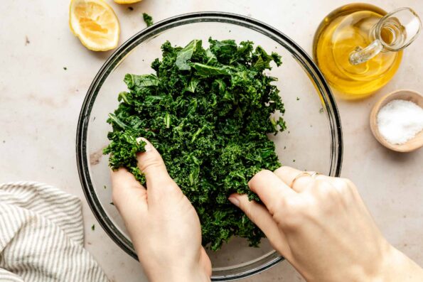 An overhead shot of a woman's hands massaging kale in a glass bowl atop an off-white surface. olive oil, salt, a striped cloth and squeezed lemon wedges sit beside the bowl.