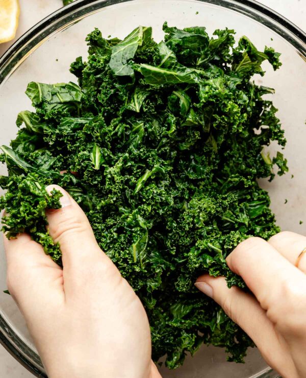 An overhead shot of a woman's hands massaging kale in a glass bowl atop an off-white surface.