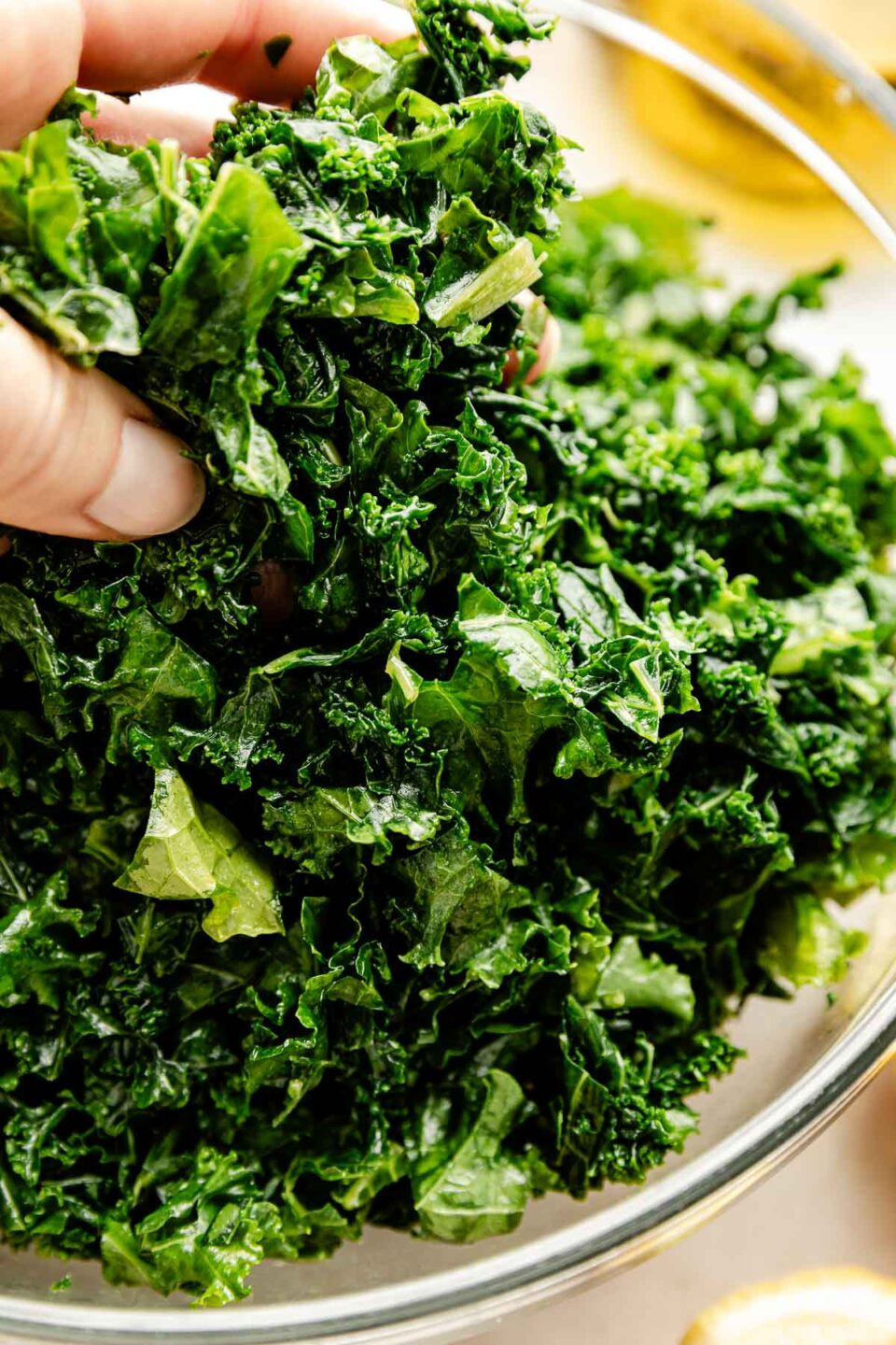 A close-up shot of a woman's hand holding up a handful of massaged kale over a glass bowl of kale.