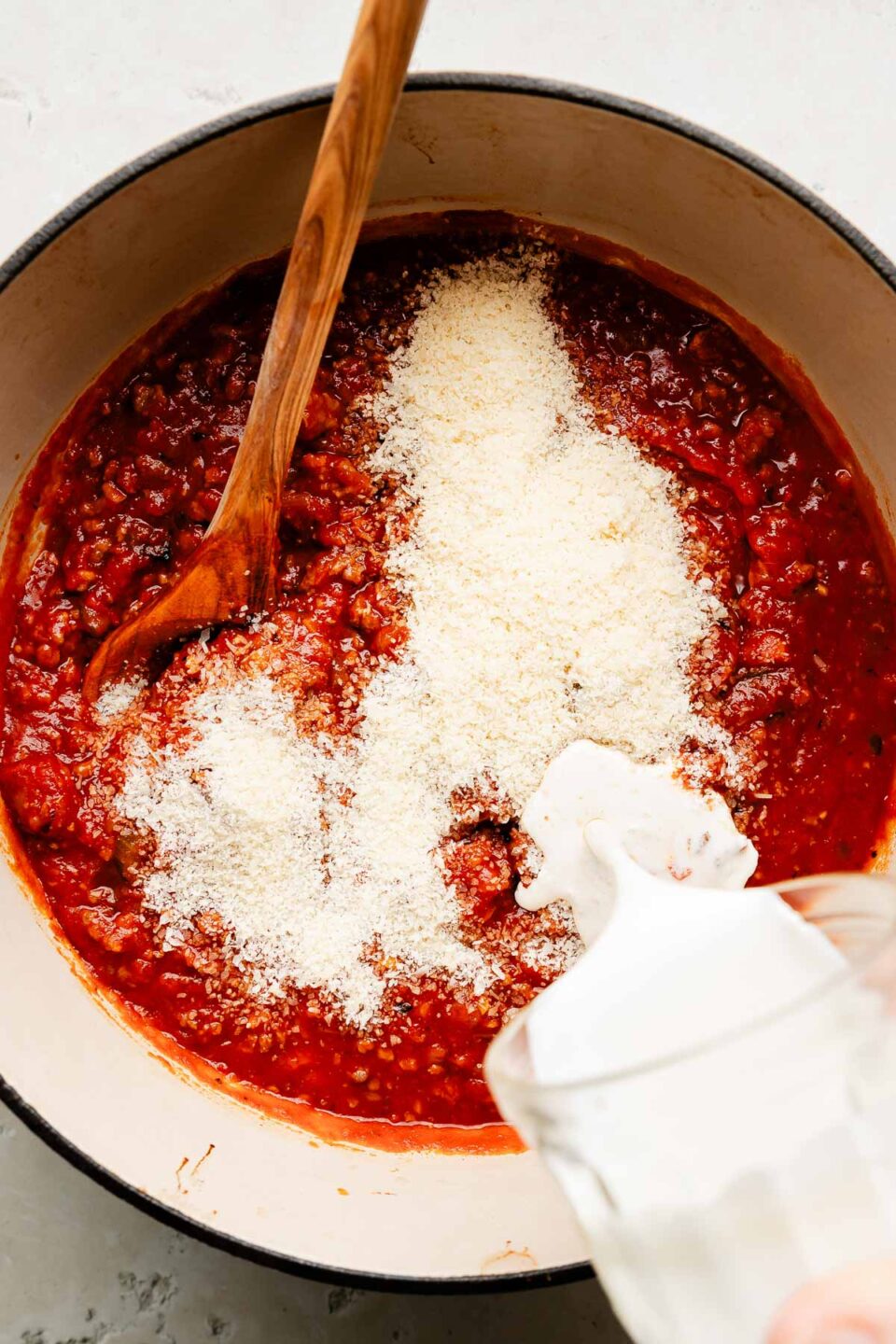 An overhead shot of heavy cream being poured into a Dutch oven of pasta sauce with grated parmesan in a Dutch oven sitting on an off-white surface.