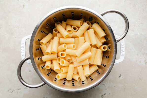 An overhead shot of cooked pasta in a colander over a white Dutch oven sitting on an off-white surface.