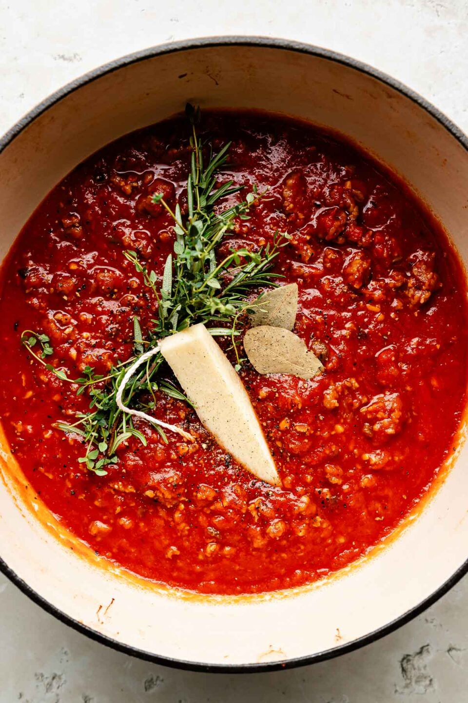 An overhead shot of sausage ragu topped with a parmesan rind, a bundle of fresh herbs, and two bay leaves in a Dutch oven atop an off-white surface.