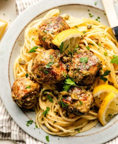 An overhead shot of a serving of chicken piccata meatballs on a bed of spaghetti noodles on a white stoneware plate atop a striped cloth on an off-white surface.
