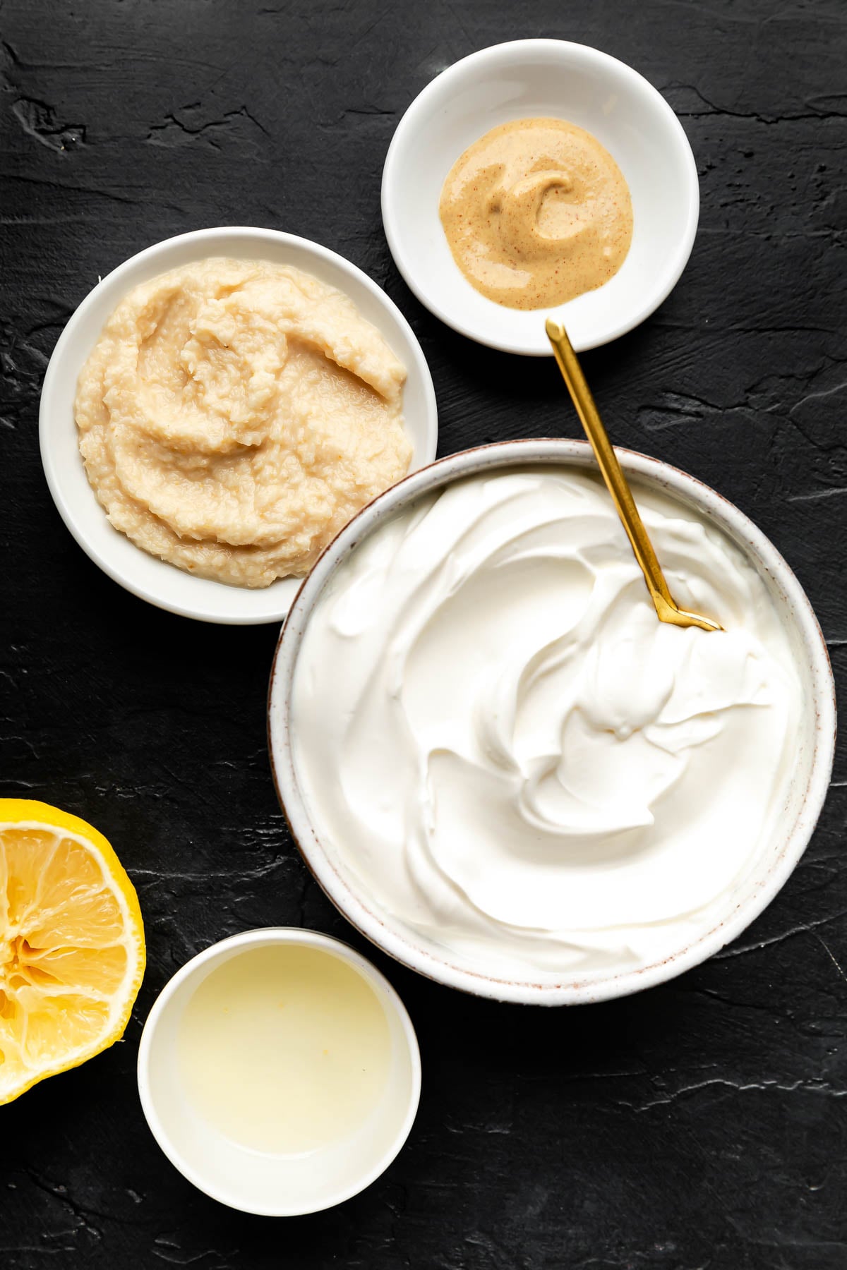 An overhead shot of ingredients in white dishes on a black surface: sour cream, horseradish, dijon mustard, and lemon juice.