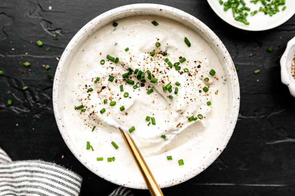 An overhead shot of mixed homemade horseradish sauce topped with black pepper and chopped chives in a stoneware bowl atop a black textured surface. A striped cloth and small dishes of chopped chives and black pepper sit alongside it.