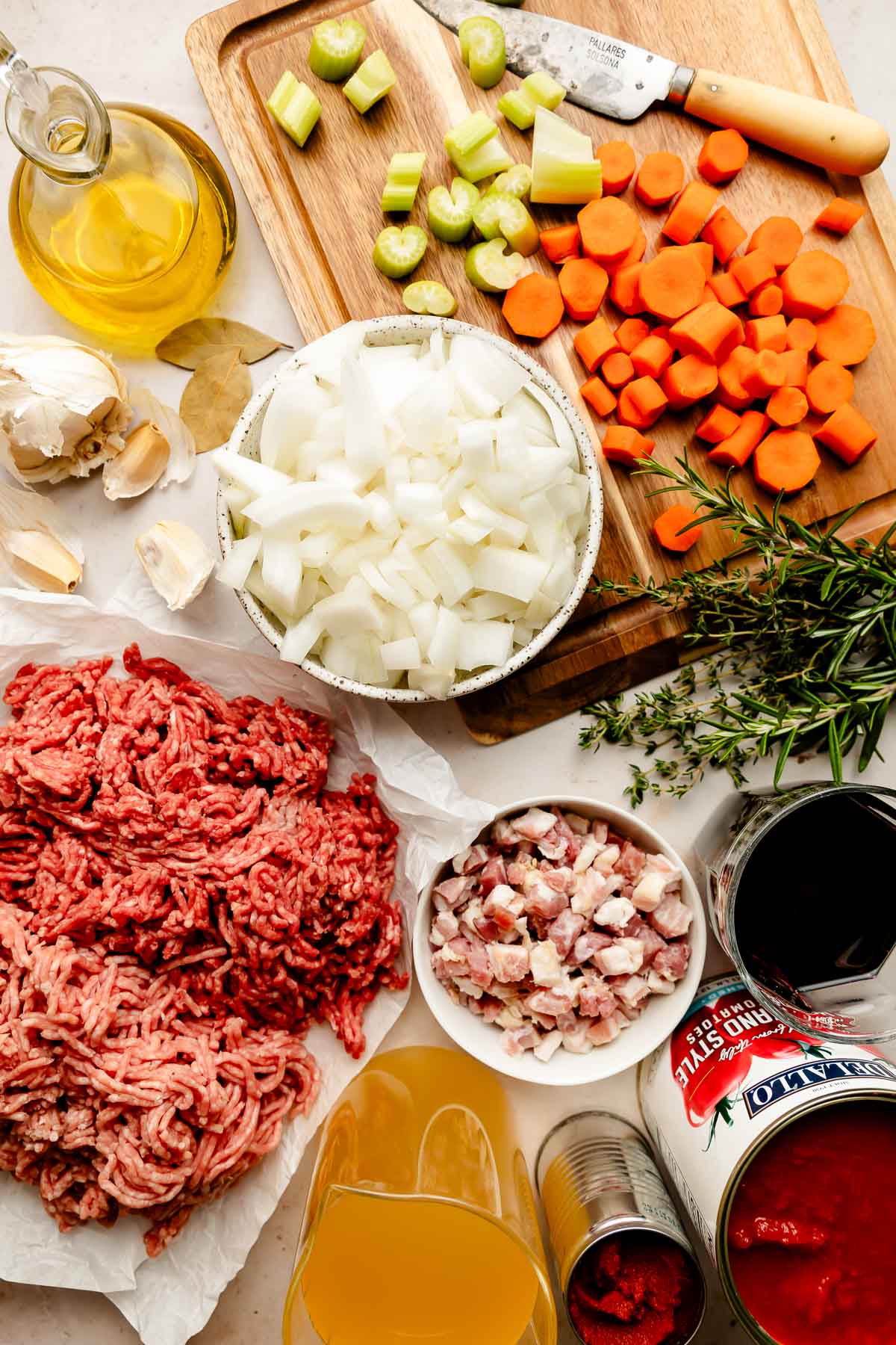 An overhead shot of ingredients displayed on a wooden cutting board and a white surface: chopped celery, carrots, and onion, pancetta, ground pork and beef, fresh herbs, garlic, olive oil, crushed tomatoes, chicken broth, and tomato paste.