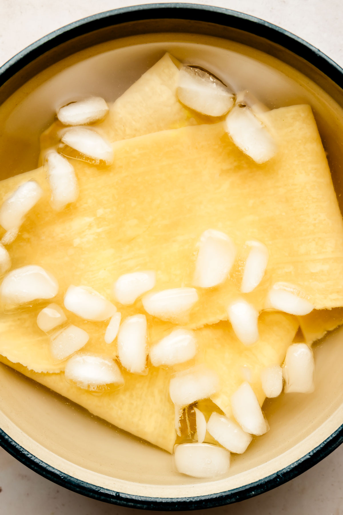 An overhead shot of fresh lasagna noodles in an ice bath in a Dutch oven atop an off-white surface.