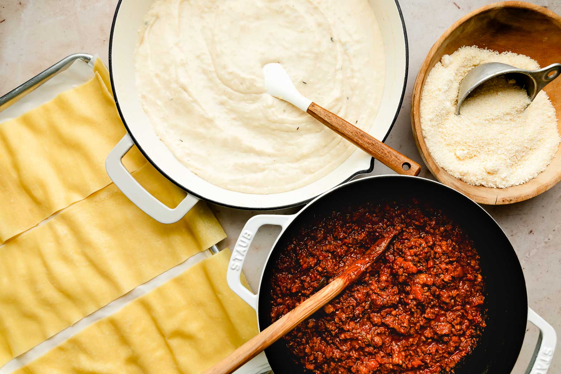 An overhead shot of a sheet pan of lasagna noodles, a skillet of bechamel sauce, a Dutch oven full of bolognese sauce and a wooden bowl of grated parmesan on an off-white surface.