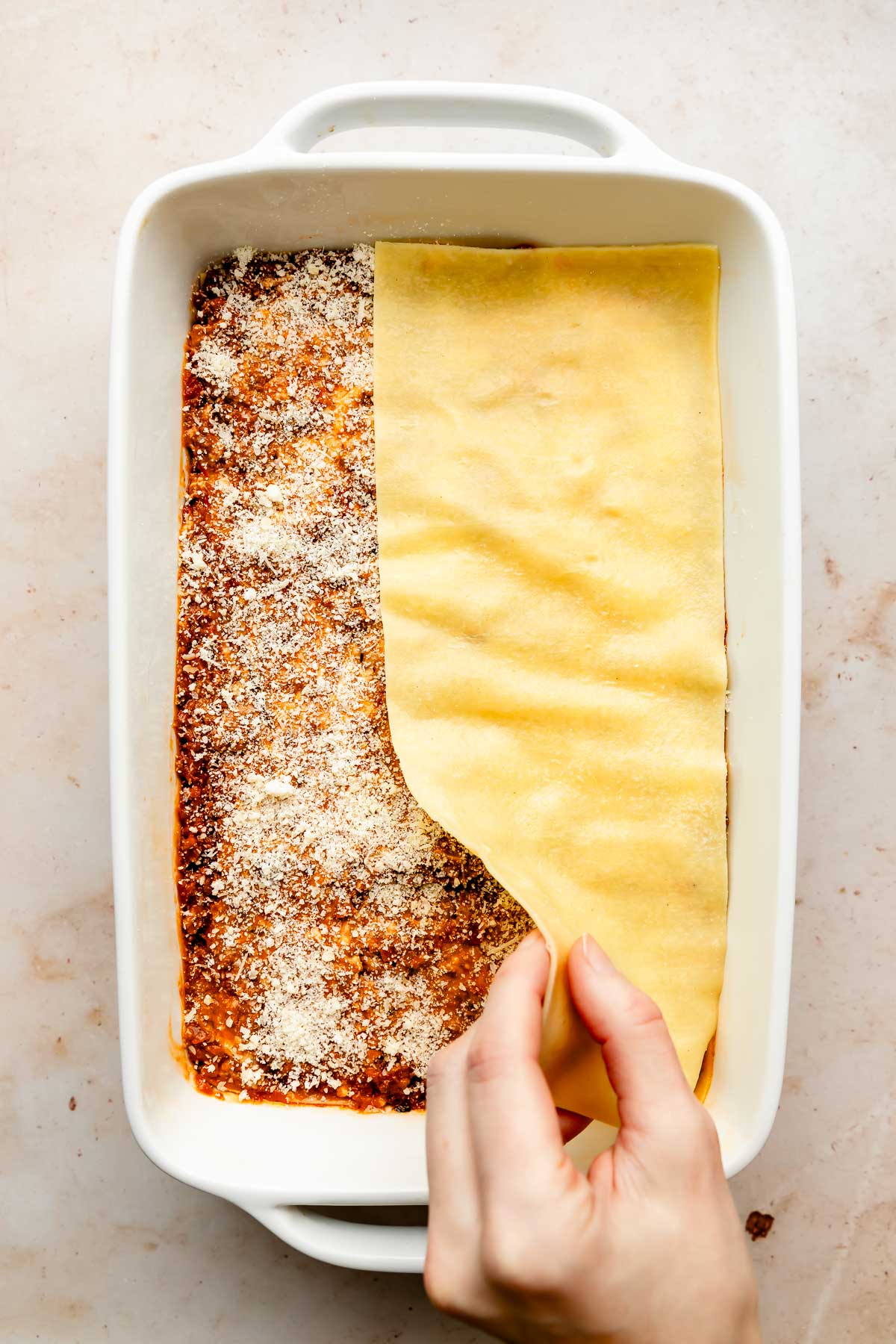 An overhead shot of a woman's hand laying a lasagna sheet over the top of bolognese sauce and parmesan in a white baking dish atop an off-white surface.