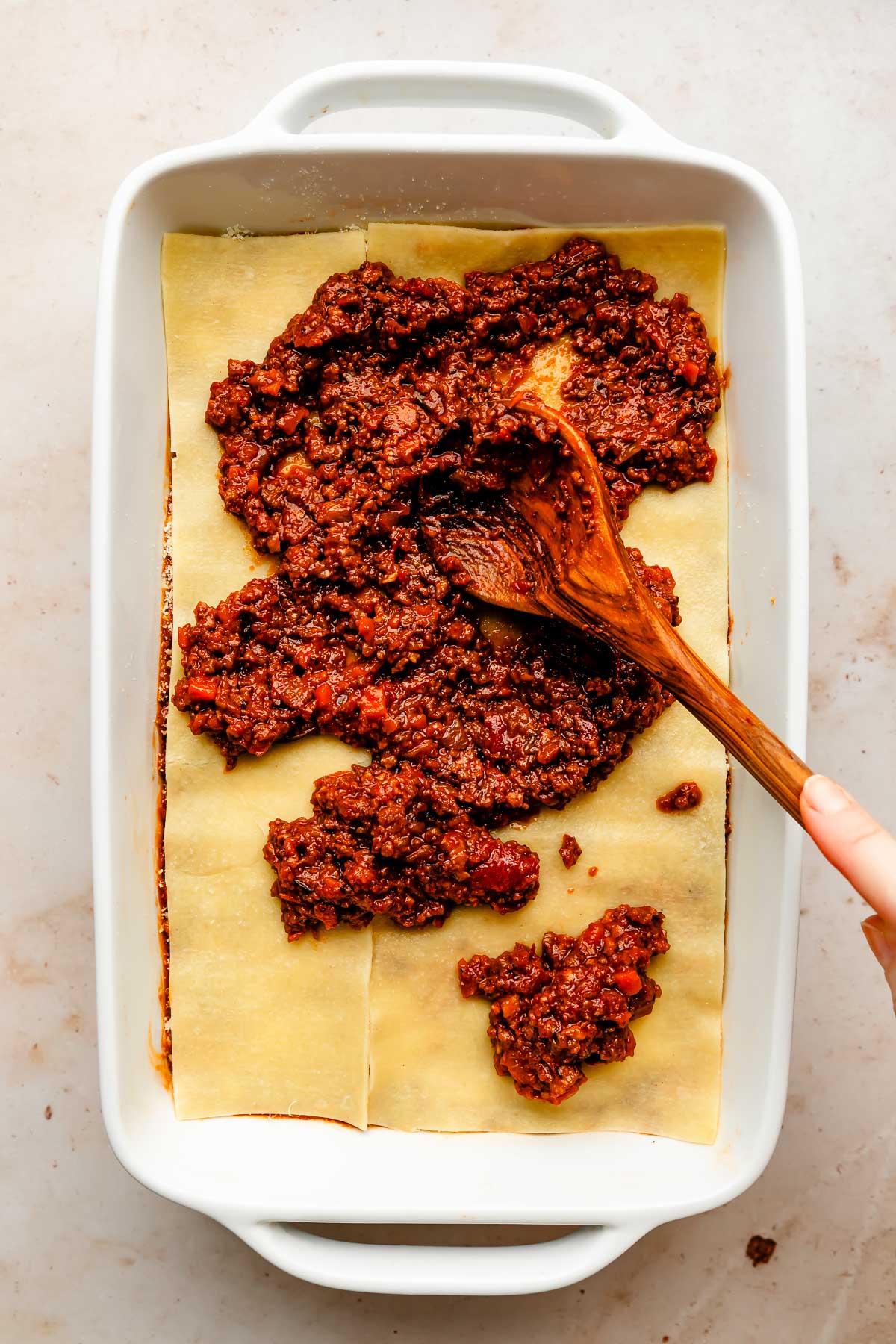An overhead shot of a wooden spoon spreading bolognese sauce over a layer of lasagna noodles in a white baking dish atop an off-white surface.