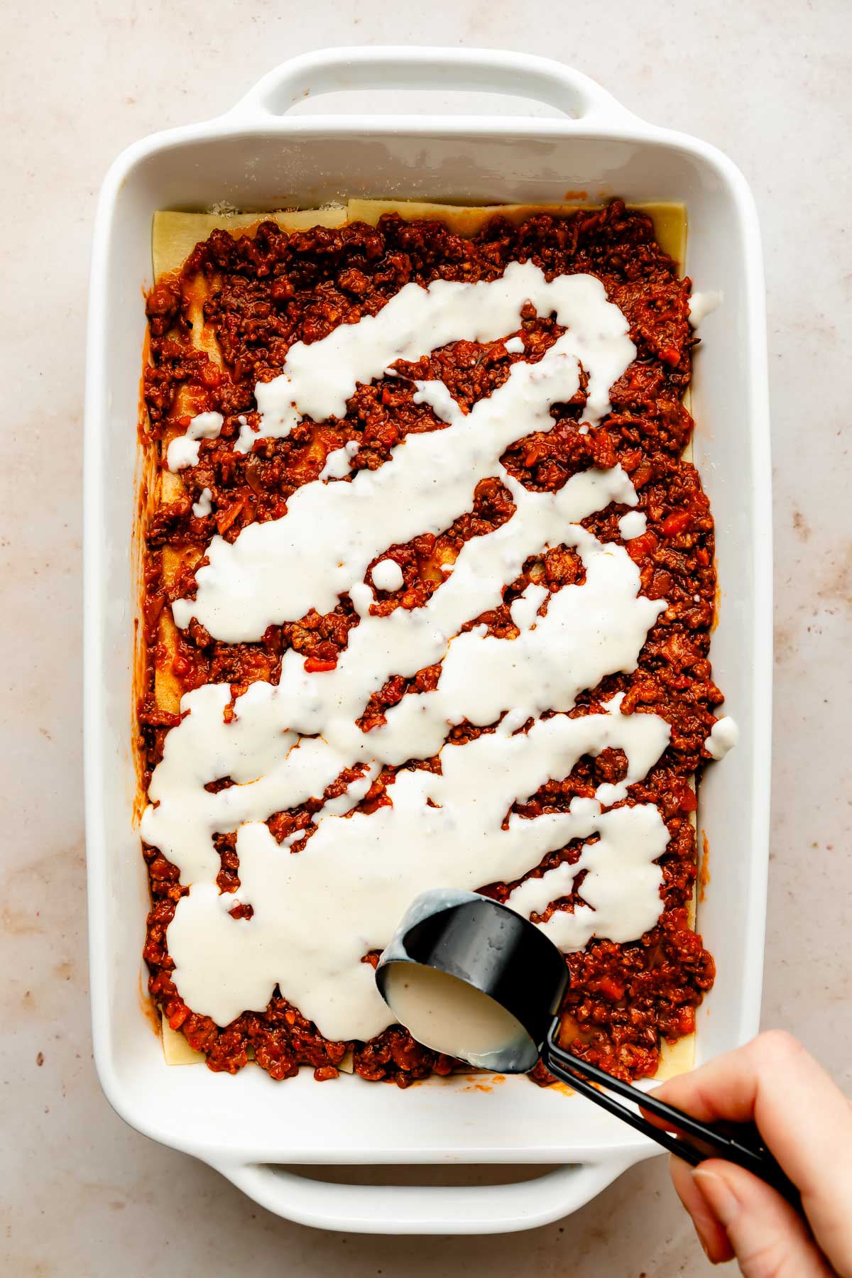 An overhead shot of a woman's hand spreading bechamel sauce over a layer of bolognese sauce in a white baking dish atop an off-white surface.
